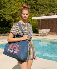 A.P.C. Sales & Events - Woman standing by pool in t-shirt and shorts with A.P.C. tote bag.