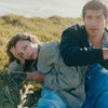 female model on the ground, wearing a green jacket and a denim. Male model next to her, wearing dark blue jacket and printed shirt.